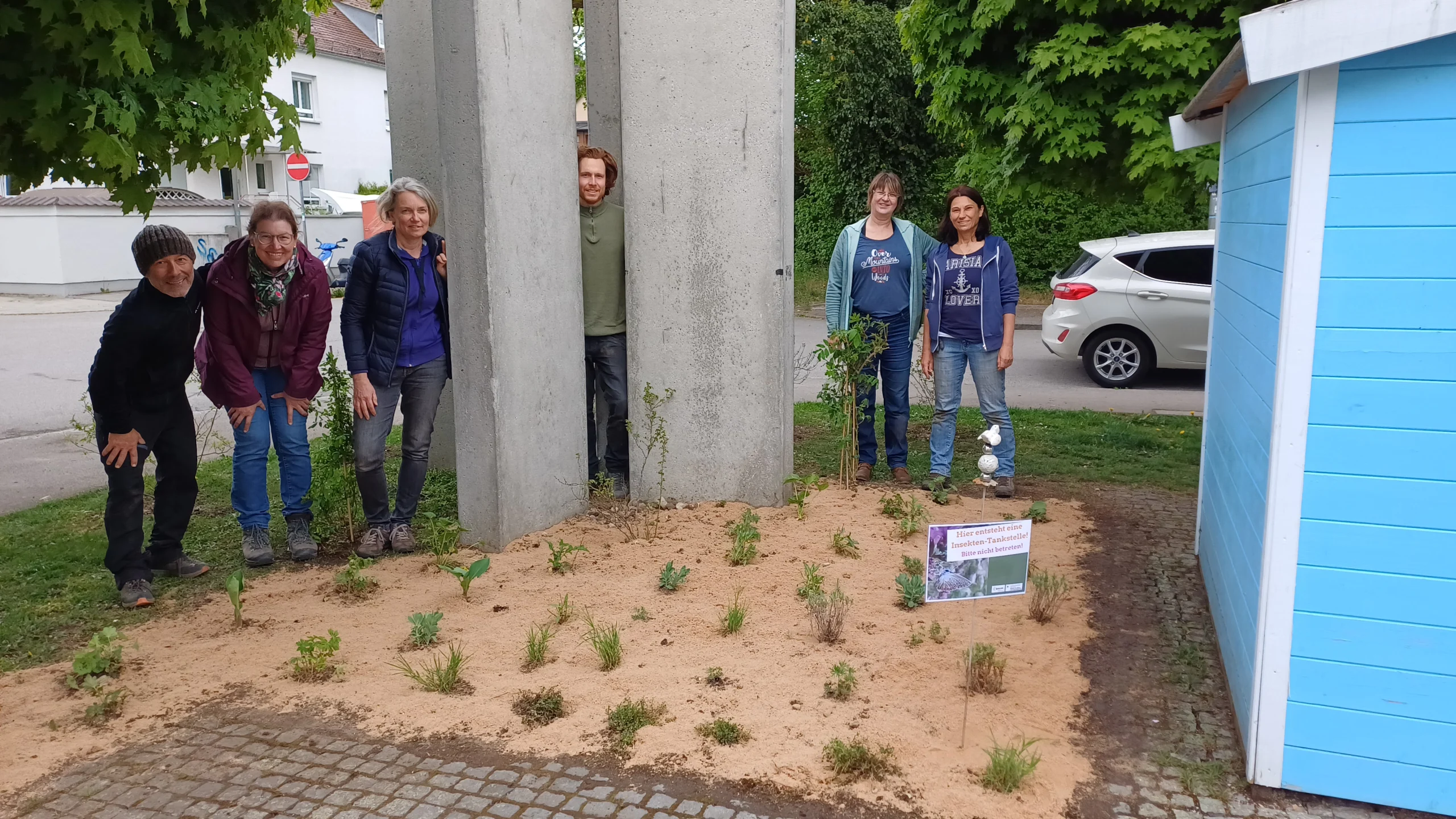 Neu angelegte Blühfläche neben einem blauen Holzhäuschen, im Hintergrund mehrere Menschen