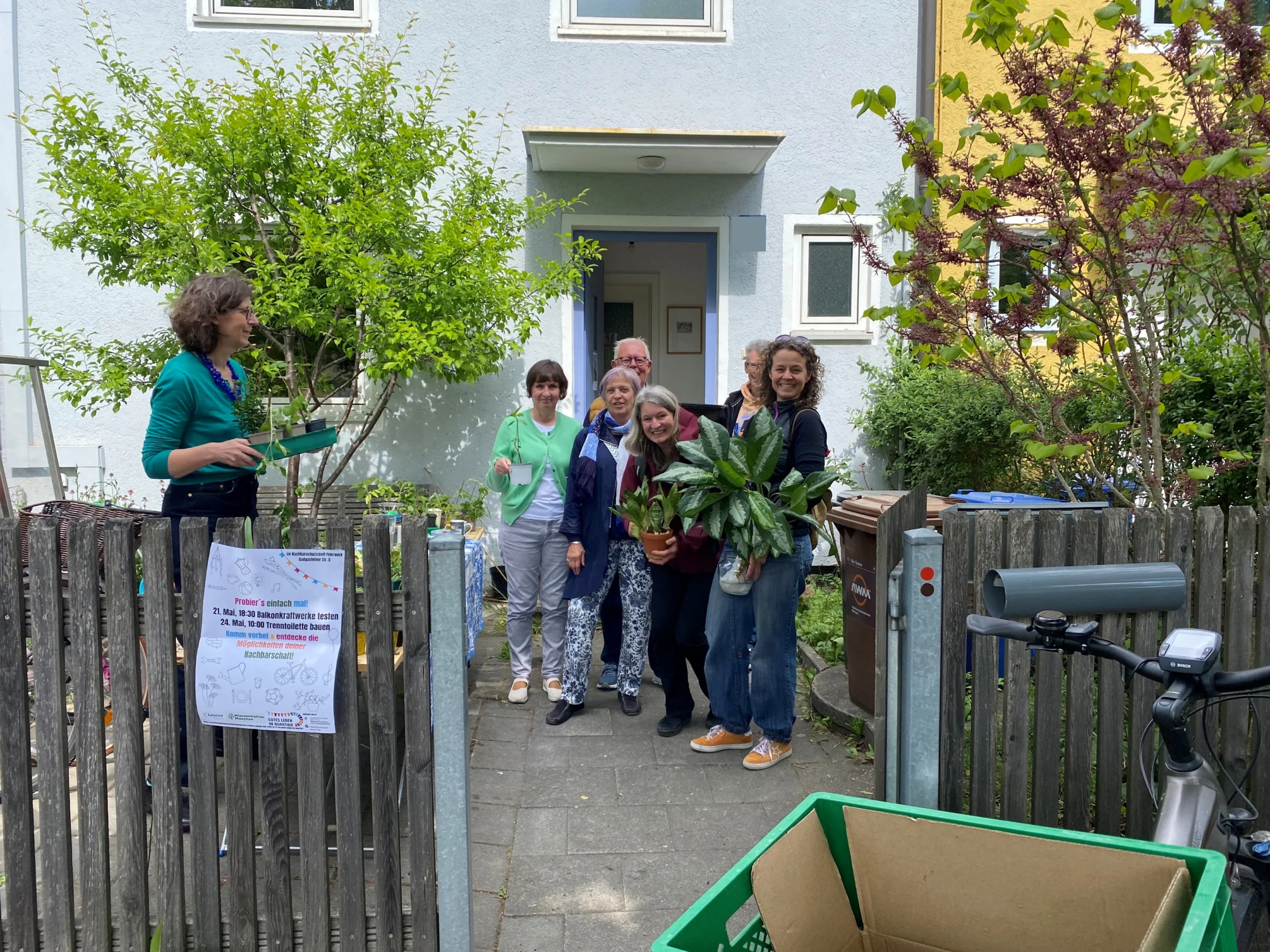 Menschen stehen mit Pflanzen vor einem Haus mit Gartenzaun